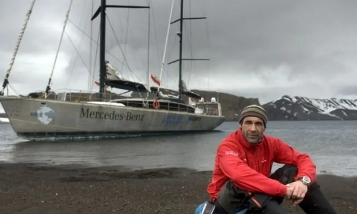 Mike Horn, l'explorateur, pose près de son bateau "Mercedes-Benz" sur une plage rocheuse, avec des montagnes enneigées en arrière-plan.