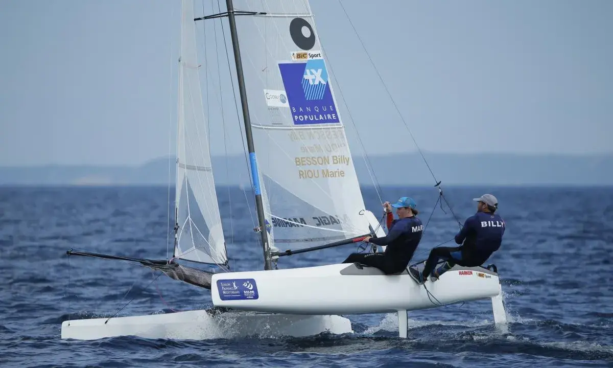 Deux marins, dont Billy Besson, naviguent sur un catamaran blanc par temps calme.