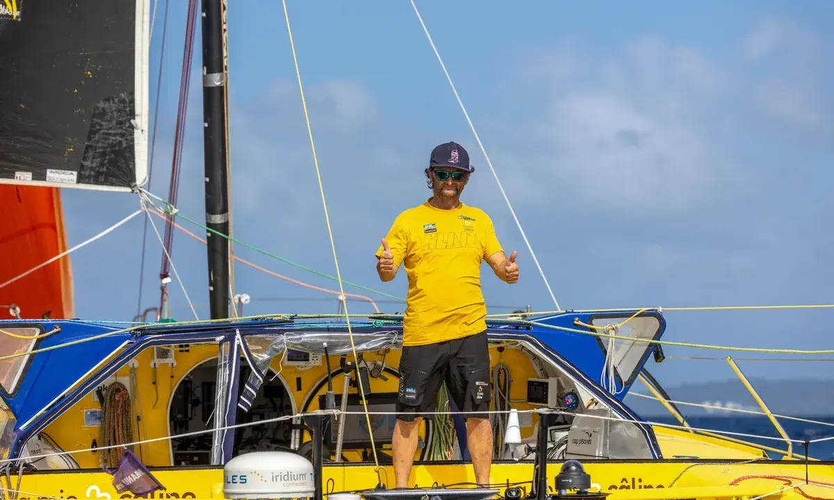 Arnaud Boissières, souriant, fait un double pouce levé sur son voilier jaune et bleu, prêt pour une nouvelle aventure en mer.