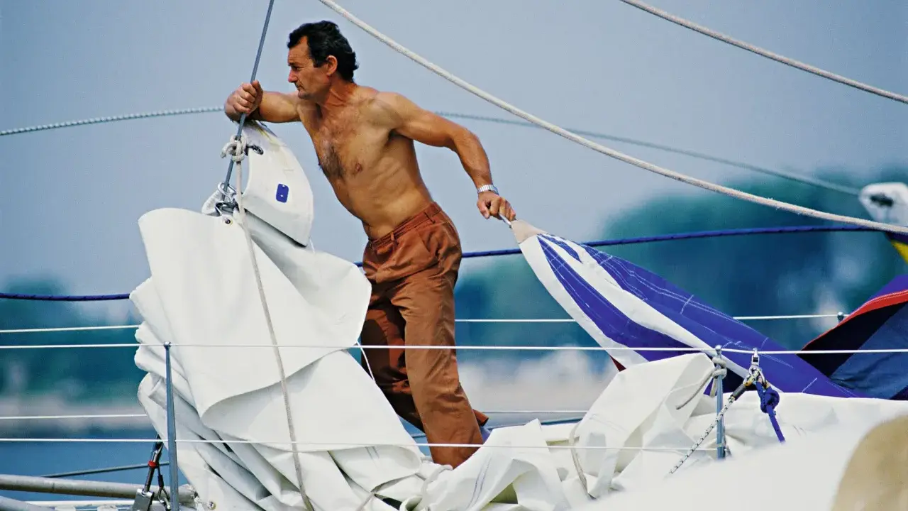 Un marinier, peut-être Eric Tabarly, manipule des voiles sur un bateau. Sa fortune est dans le vent et l'océan.