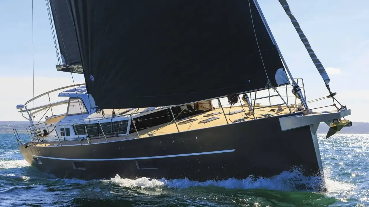 Un voilier moderne, avec une grande voile noire, navigue sur une mer agitée. Xavier Desmarest admire la vue depuis le pont.