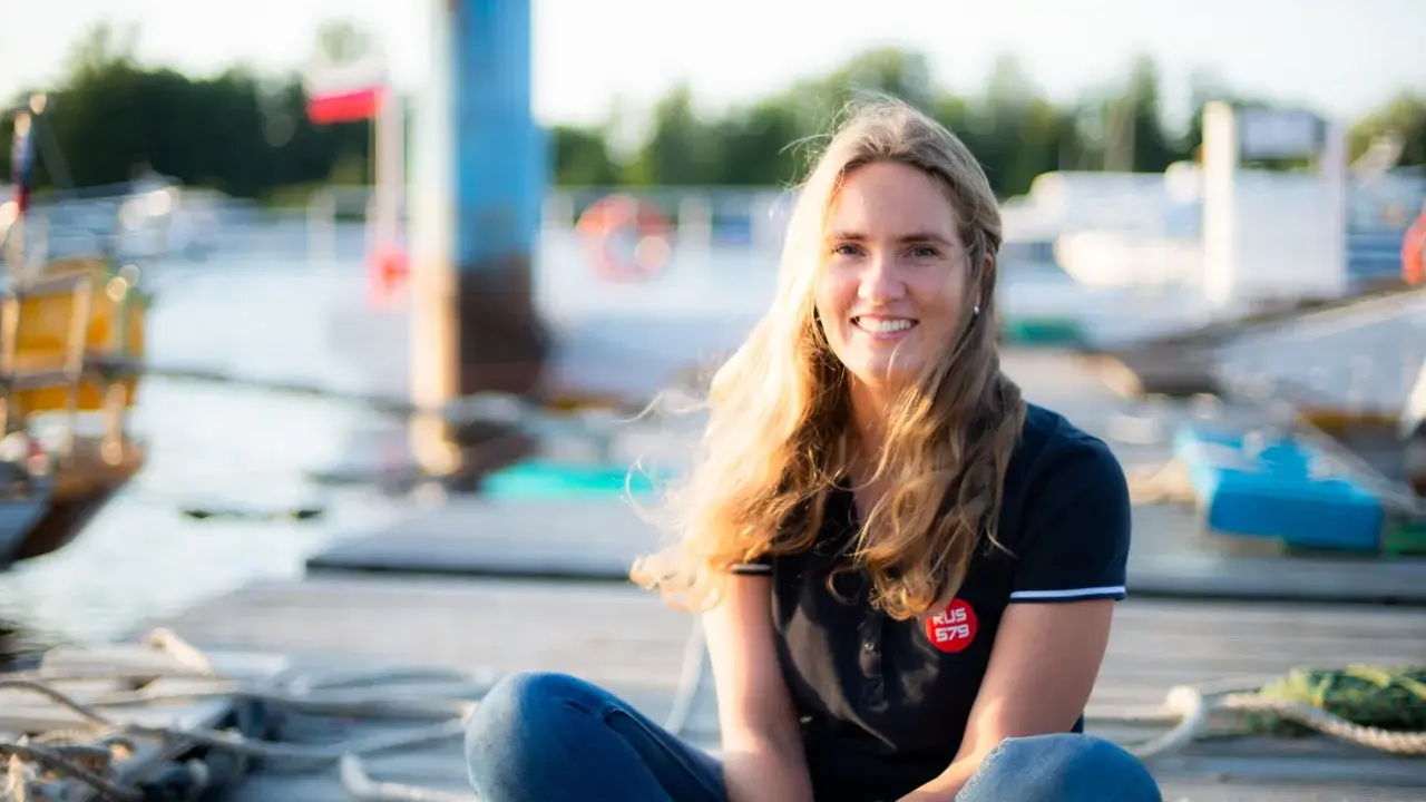 Irina Gracheva, souriante, assise sur un ponton en bois, avec des bateaux et des bouées en arrière-plan.