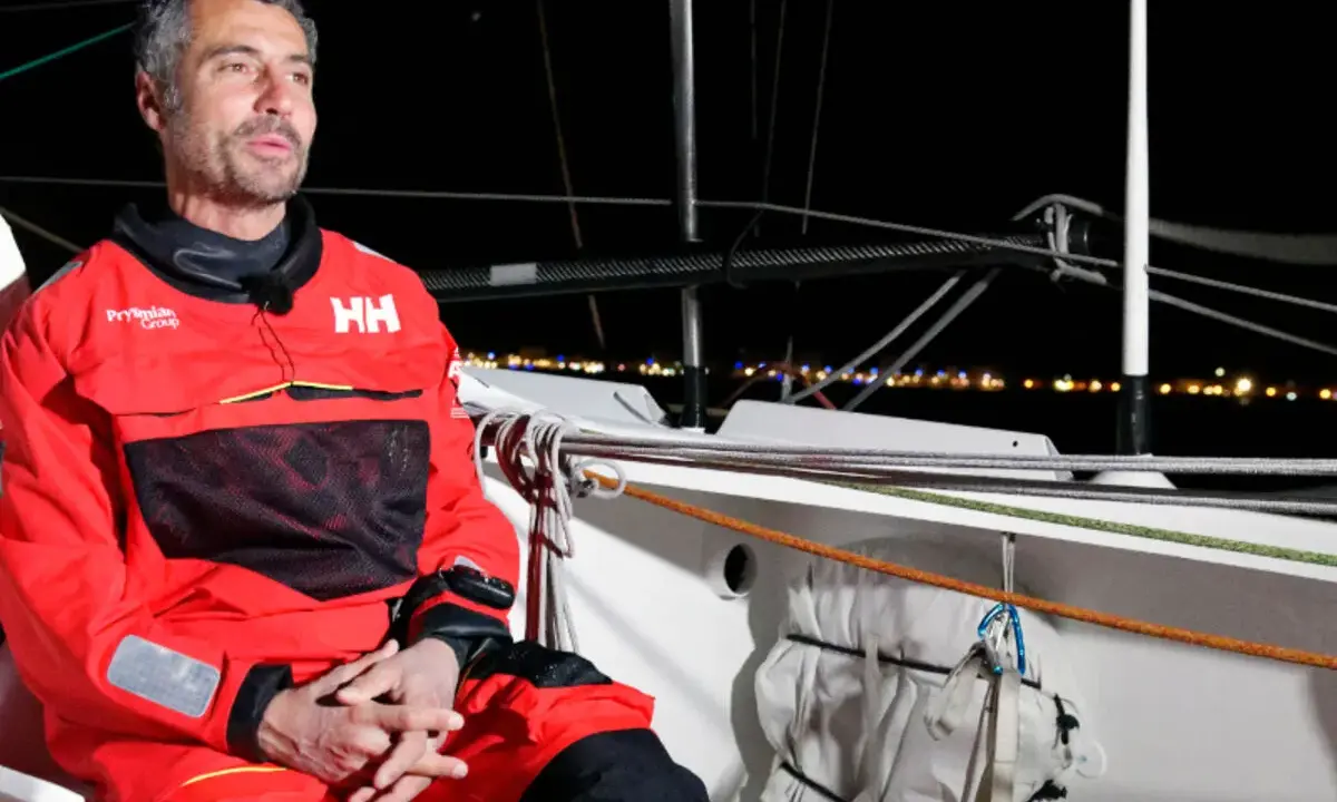 Giancarlo Pedote, skipper en tenue de voile rouge, sur un bateau la nuit, avec des lumières de ville en arrière-plan.