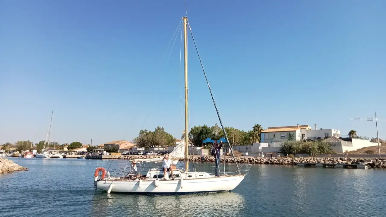 Un voilier atlante navigue sur des eaux calmes, avec des personnes à bord. Le ciel est bleu et dégagé.