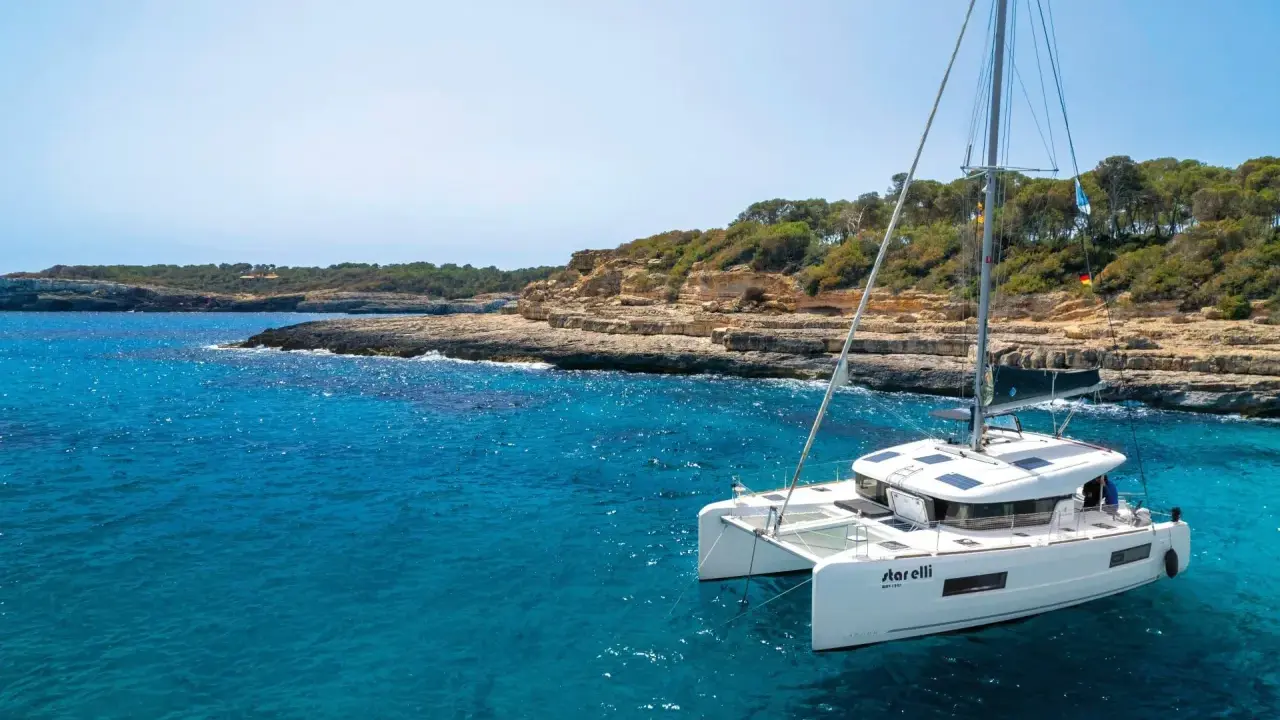 Un catamaran blanc, le "StarElli", navigue dans des eaux turquoise près d'une côte rocheuse et verdoyante. Ce safran catamaran offre une vue idyllique.