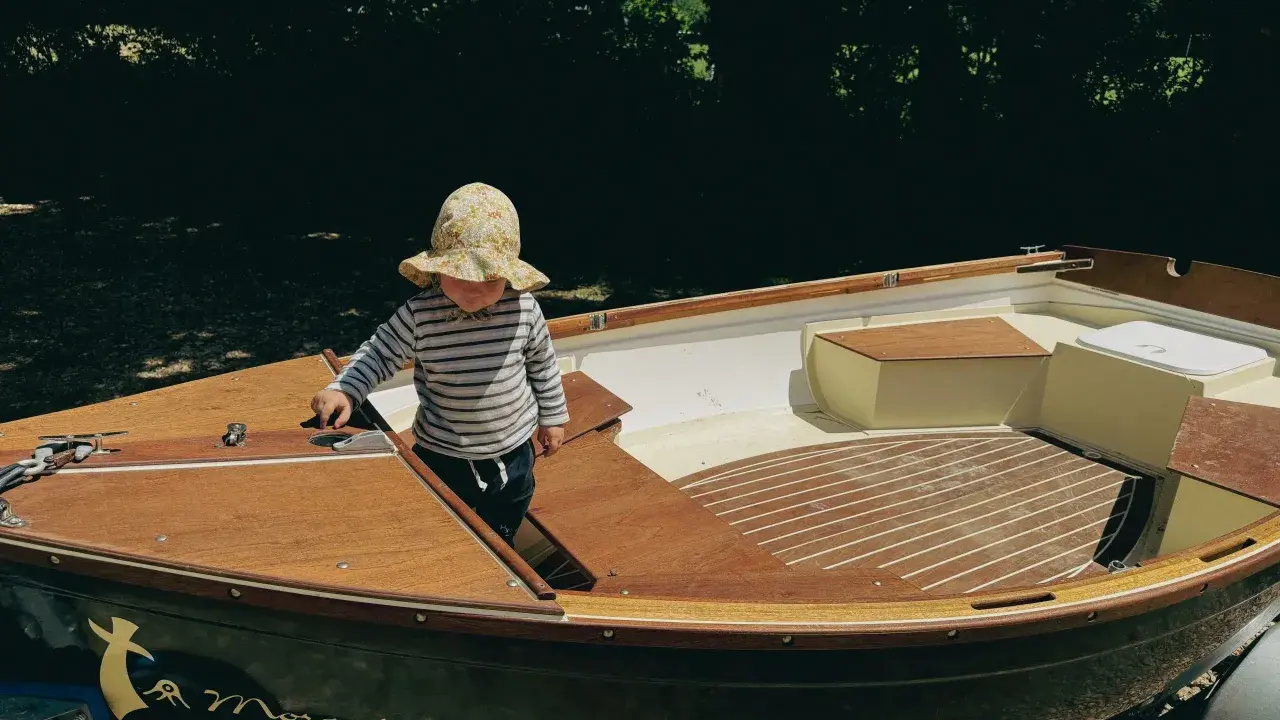 Un enfant en chapeau fleuri explore l'intérieur d'un bateau en bois, le Morgat 320, prêt pour l'aventure.
