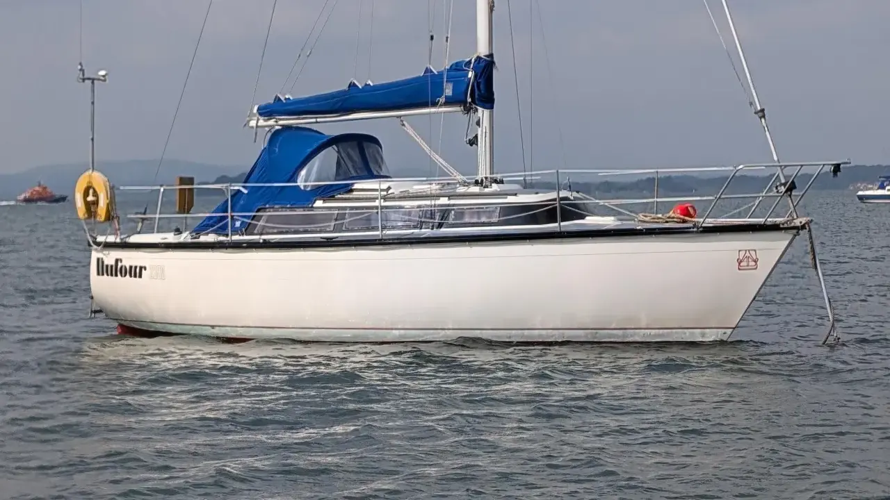 Voilier blanc Dufour 2800 à l'ancre, avec une bâche bleue sur la cabine. Un remorqueur orange est visible au loin.