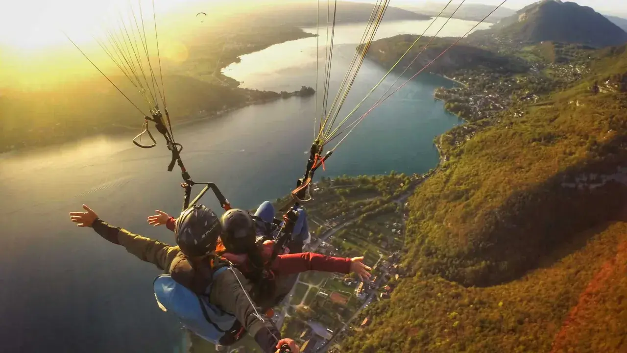 Vol en parapente biplace au-dessus d'un lac, avec un paysage de montagne et de village. Philippe Jeantot, malgré un accident passé, continue de voler.