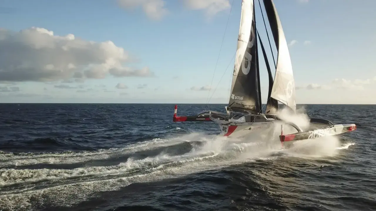 Le trimaran "Actual" d'Yves Le Blevec fend les vagues, propulsé par le vent sous un ciel bleu.
