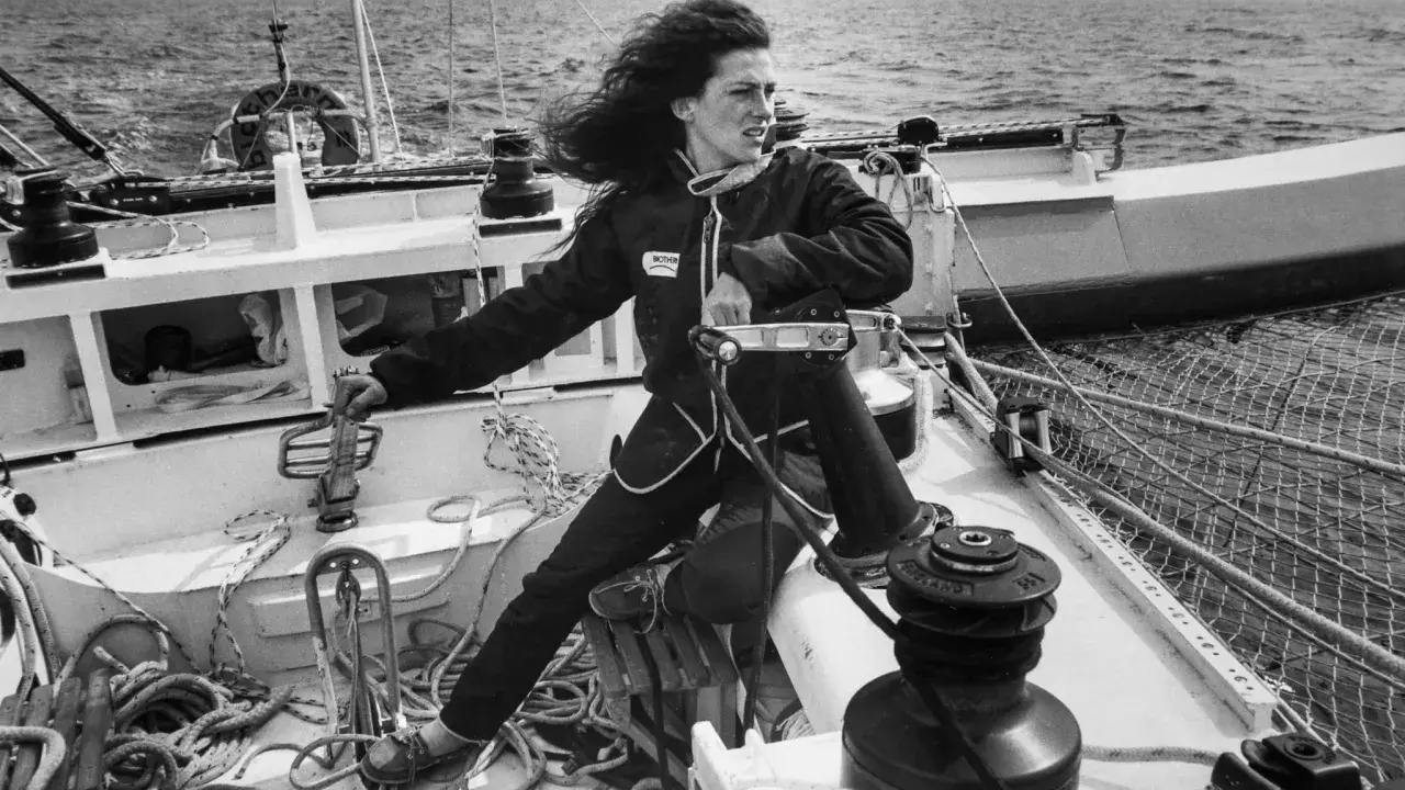 Une jeune femme aux cheveux longs, vêtue d'une combinaison de marin, manœuvre un winch sur un voilier. L'image évoque l'esprit d'aventure de Jacques Arthaud.