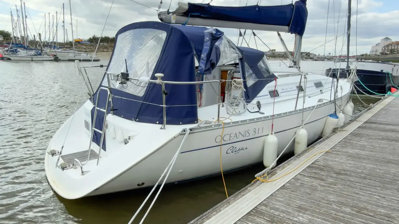 Un voilier blanc, un Océanis 311 Clipper, est amarré à un quai en bois. Des pare-battages blancs protègent sa coque.