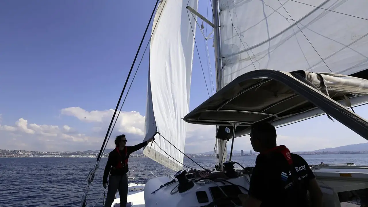 Deux hommes sur un catamaran, l'un ajuste la voile pour se mettre à la cape, tandis que l'autre tient la barre. Le soleil brille sur la mer bleue.