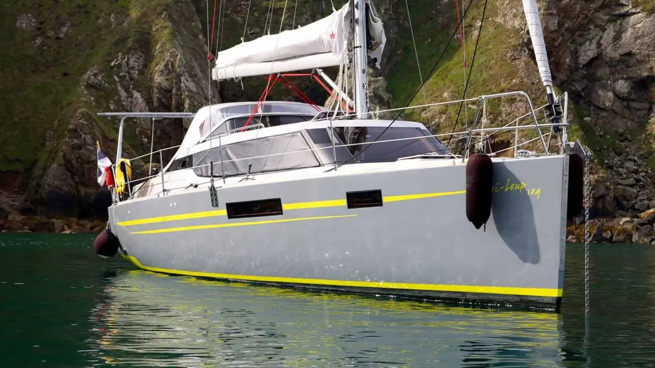 Le voilier "Bi-loup 109" gris avec des bandes jaunes, ancré près d'une falaise verdoyante.