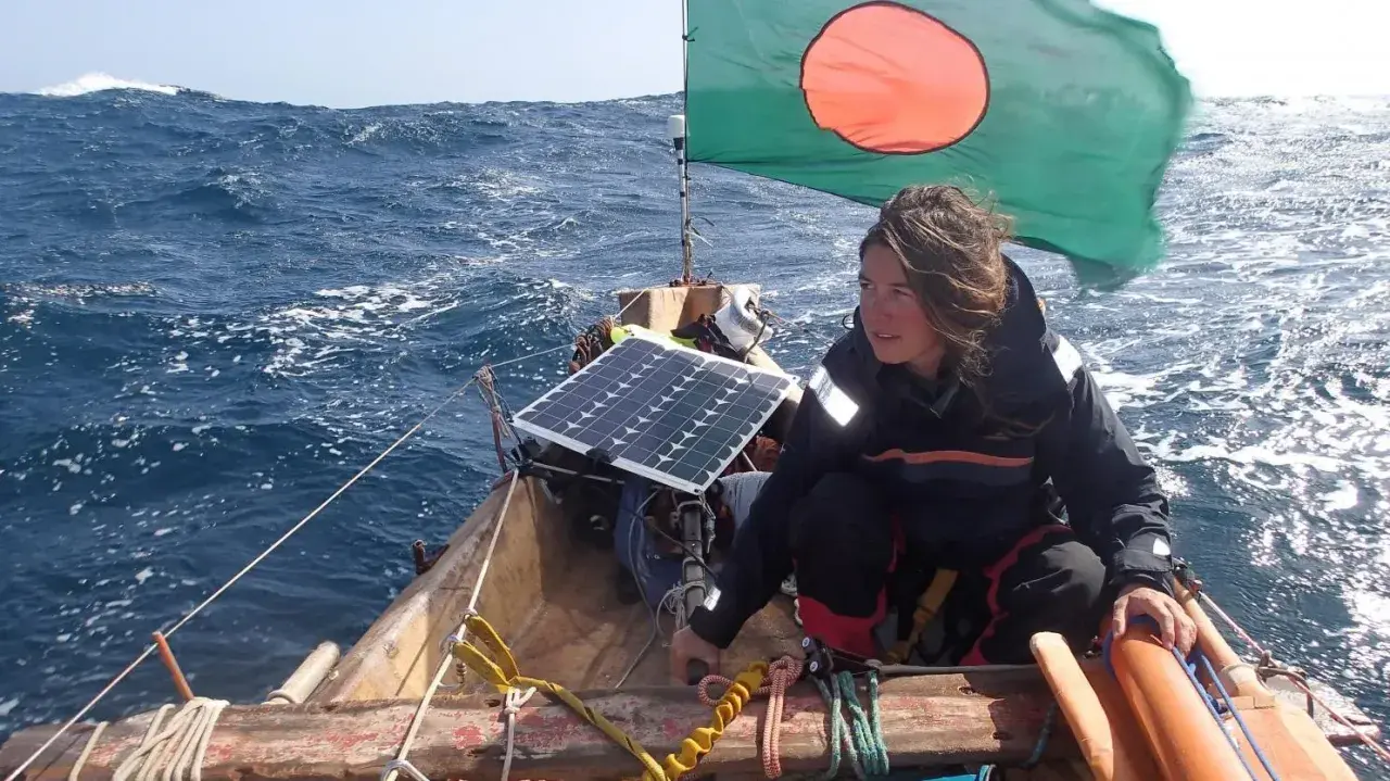 Capucine Trochet navigue sur une mer agitée, le drapeau du Bangladesh flottant au vent. Un panneau solaire est visible sur le bateau.