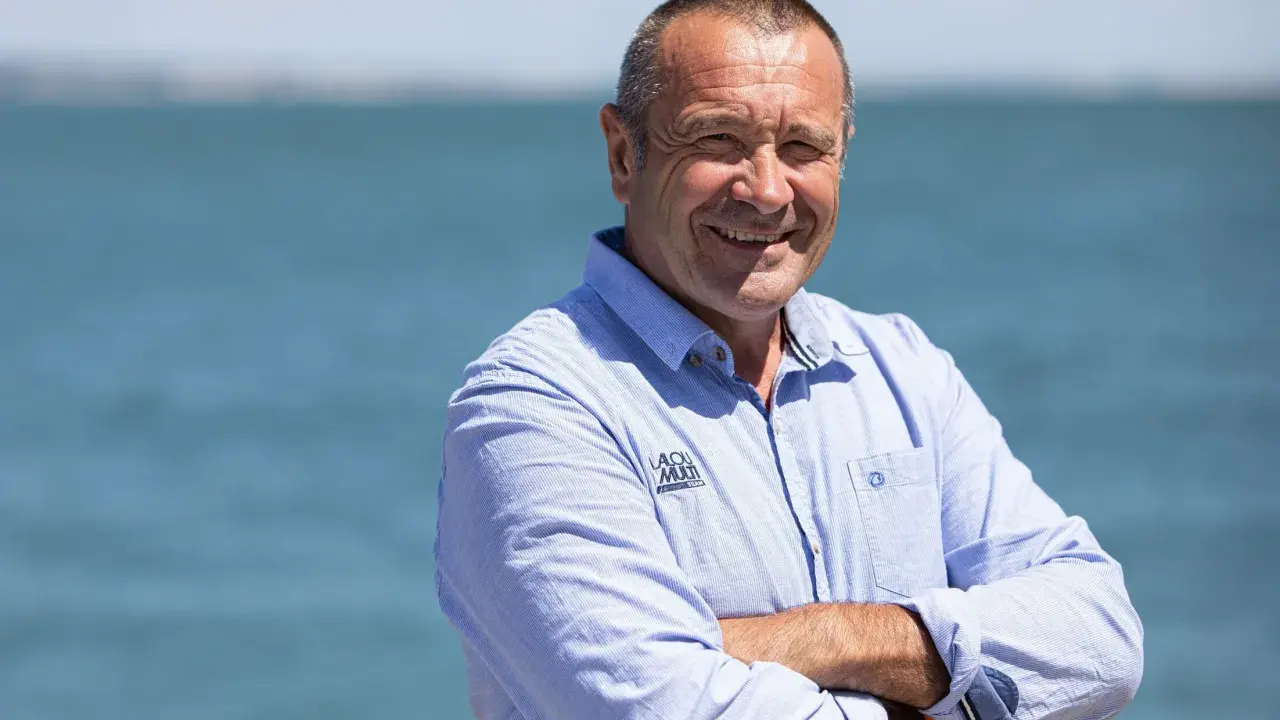 Lalou Roucayrol, souriant, bras croisés, devant la mer. Il porte une chemise bleue rayée avec un logo sur la poitrine.