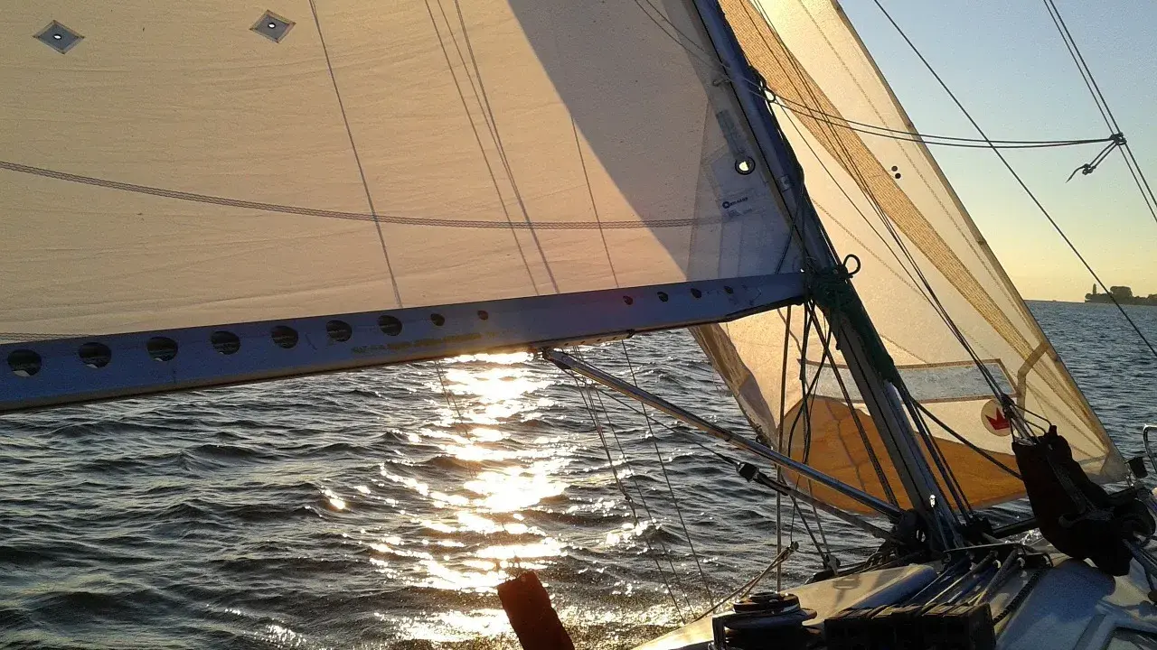 Voile d'un bateau au coucher du soleil, reflets dorés sur l'eau. La balancine du bateau est bien tendue.