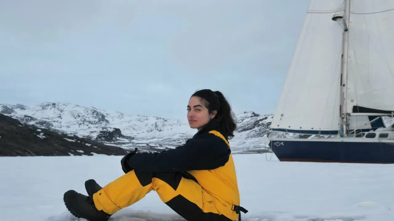 Tamara Klink, en tenue de ski jaune et noire, assise sur la neige, avec un voilier et des montagnes enneigées en arrière-plan.