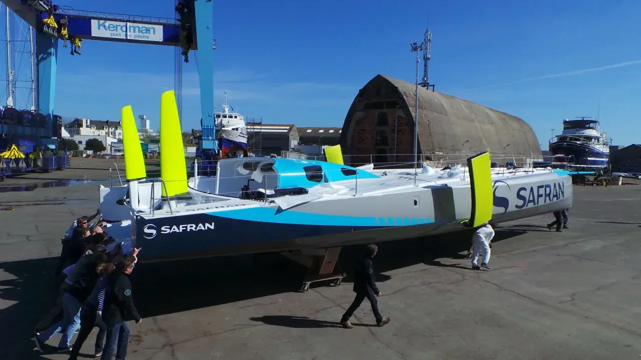 Équipe poussant un **60 pieds IMOCA** de course au large, le Safran, dans un chantier naval.