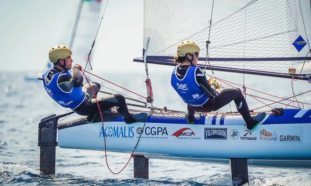 Deux jeunes navigateurs, dont Lou Berthomieu, en action sur un catamaran de sport, le vent gonflant les voiles.