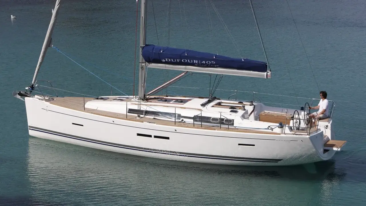 Un voilier blanc Dufour 405 Grand Large navigue sur une eau turquoise. Un homme est au poste de barre.