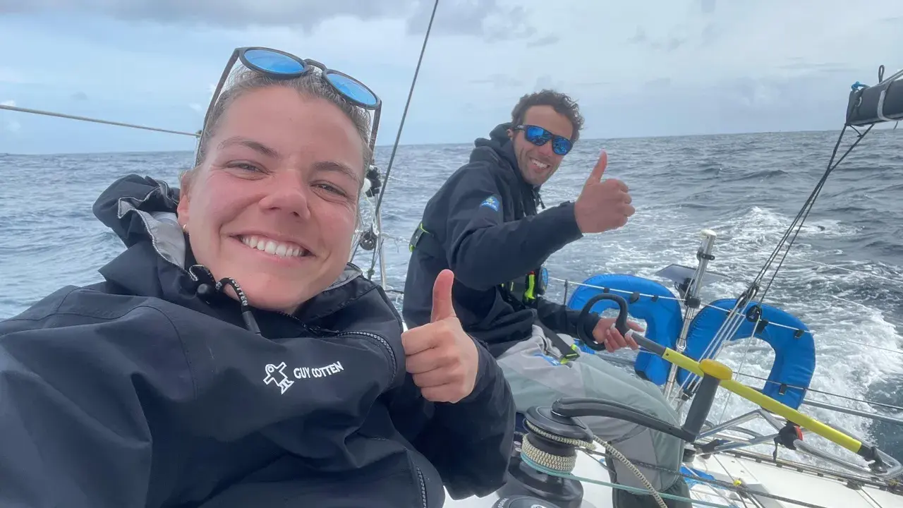 Hugo Dhallenne et sa coéquipière souriants sur un voilier, pouces levés, en pleine mer sous un ciel couvert.