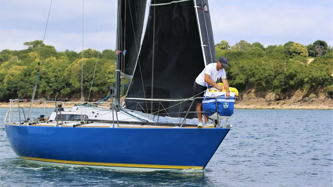 Un homme prépare le bateau, une voile noire déployée, sur fond de côte boisée. Il semble être le **half toner** de l'équipage.