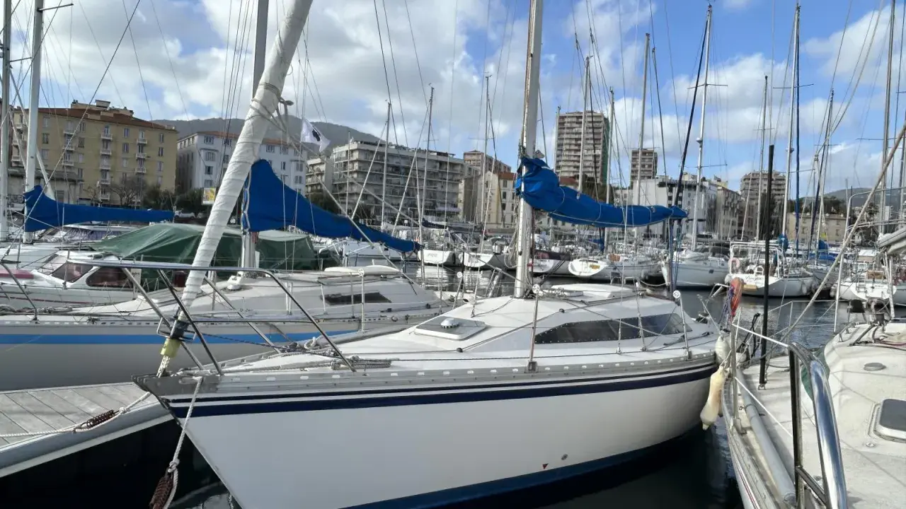Un voilier blanc aux voiles bleues, amarré dans un port. L'avis sur ce voilier rush est qu'il est prêt pour la prochaine aventure.