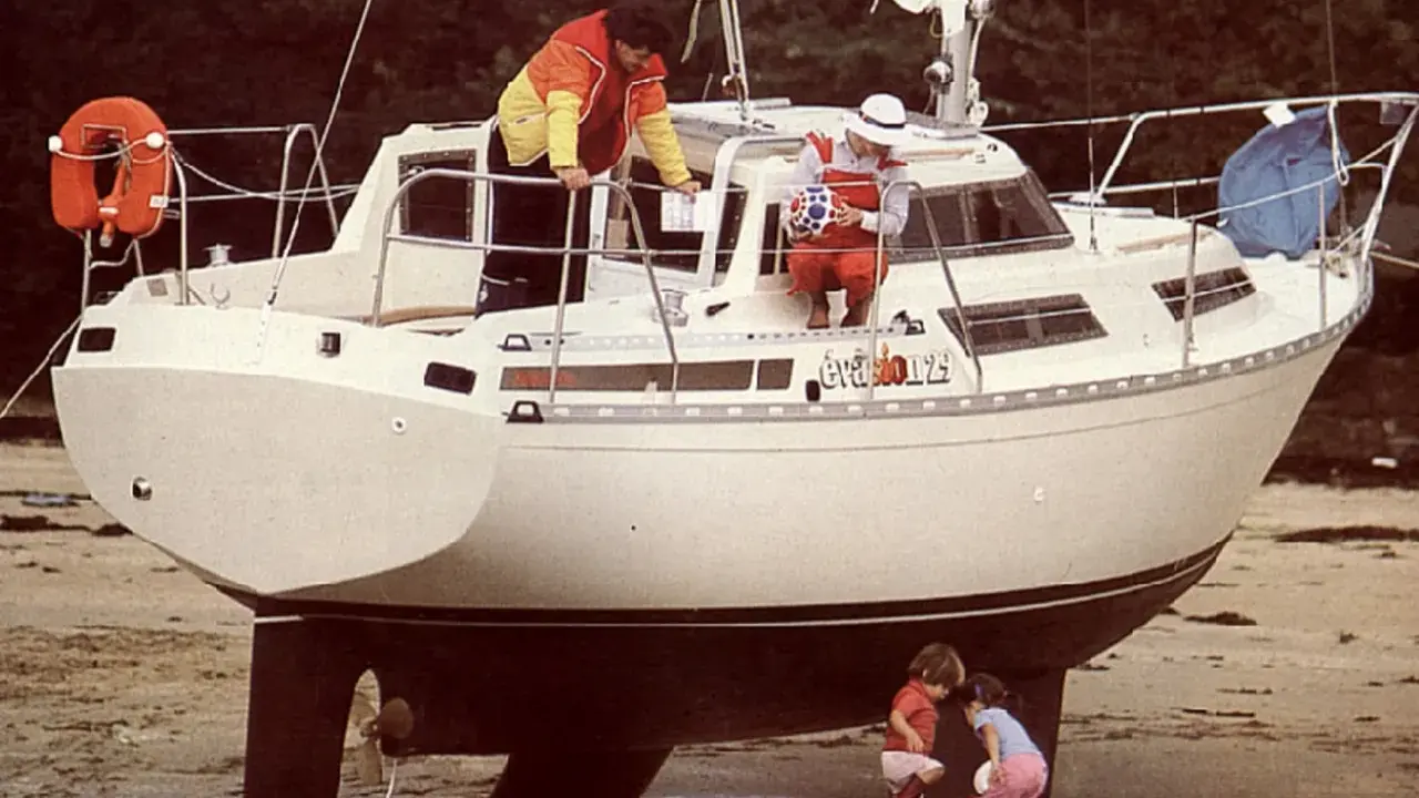 Le voilier "Évasion 29" échoué sur la plage, avec deux enfants jouant à ses pieds et deux adultes à bord.
