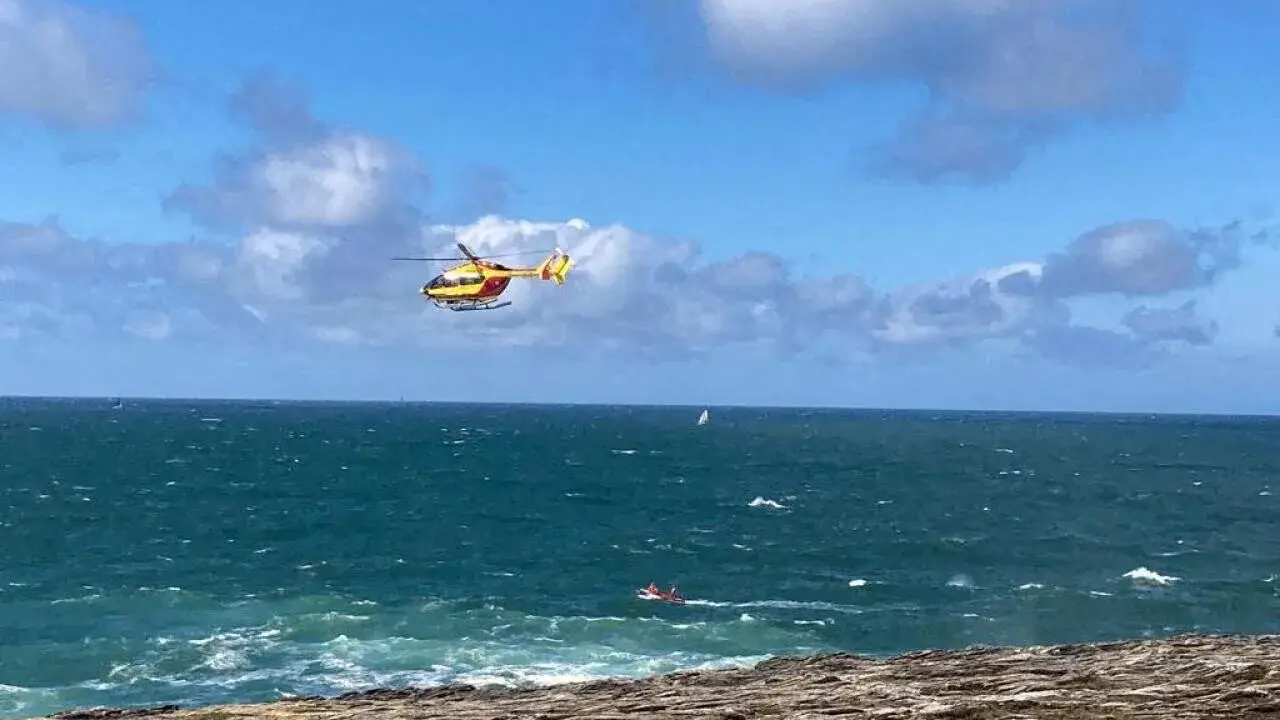 Un hélicoptère jaune et rouge survole une mer agitée, cherchant une personne disparue en mer. Un petit bateau rouge est visible au loin.