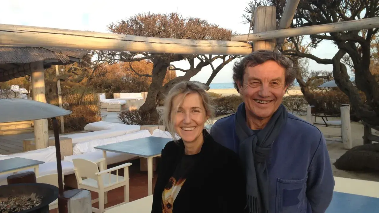 Patrice de Colmont et une femme souriants sur une terrasse de plage, avec des arbres dénudés et des sièges blancs en arrière-plan.