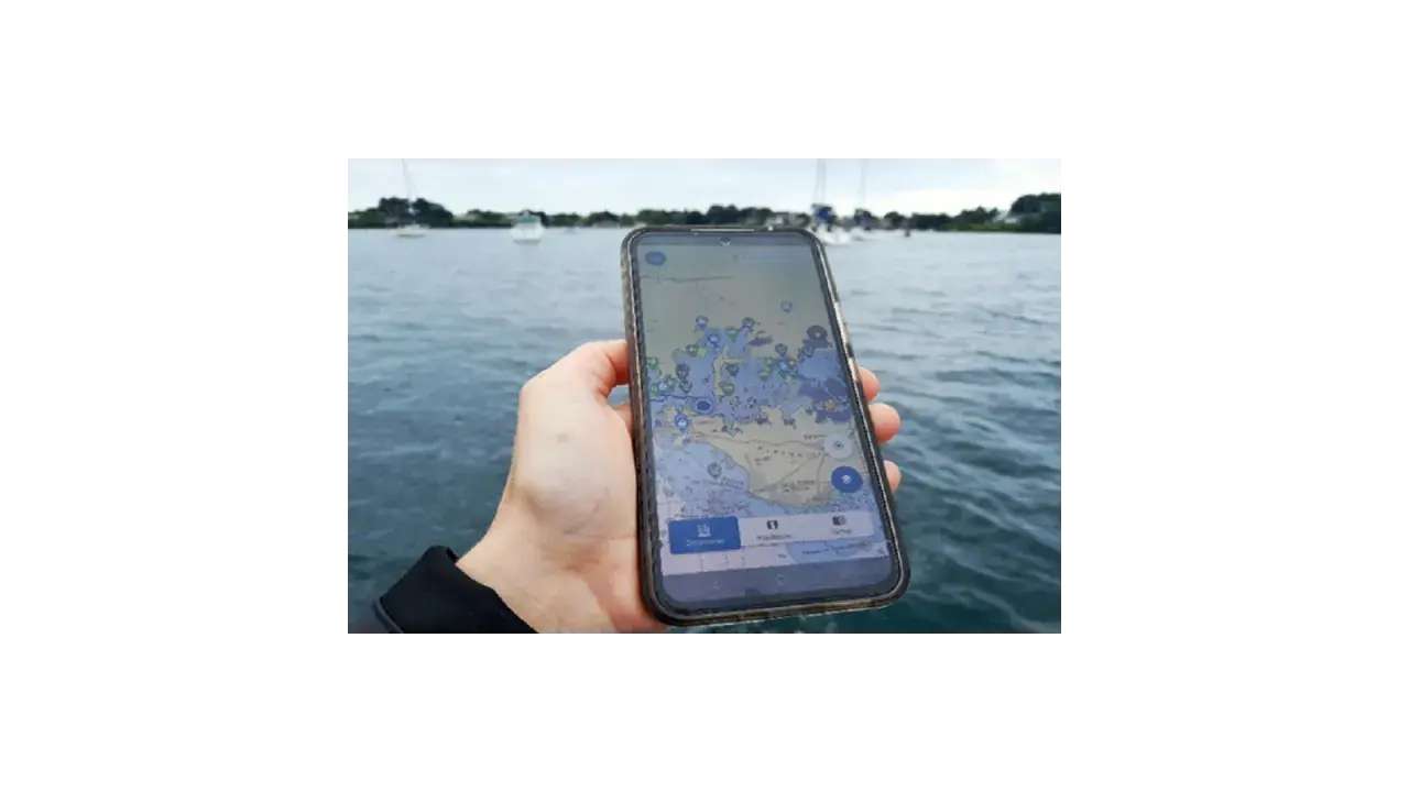 Main d'une personne tenant un smartphone affichant une carte marine avec des points d'intérêt. Le nav et co. sont visibles sur l'eau.