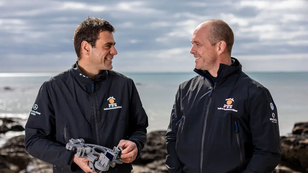 Damien Guillou et son collègue souriants sur la plage, l'un tenant un sextant, prêts pour une nouvelle aventure maritime.