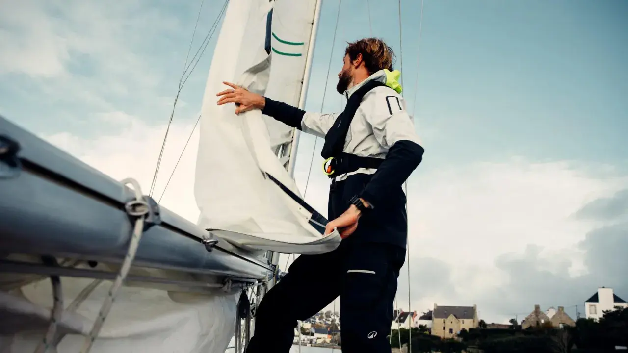 Un homme ajuste un type de voile sur un voilier, avec des maisons en arrière-plan.