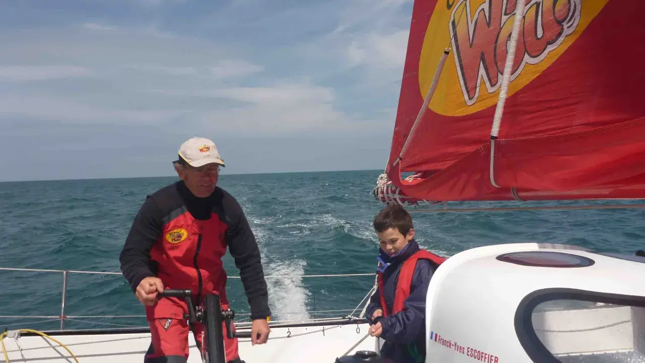 Franck-Yves Escoffier et un jeune garçon naviguent sur un voilier par temps ensoleillé.
