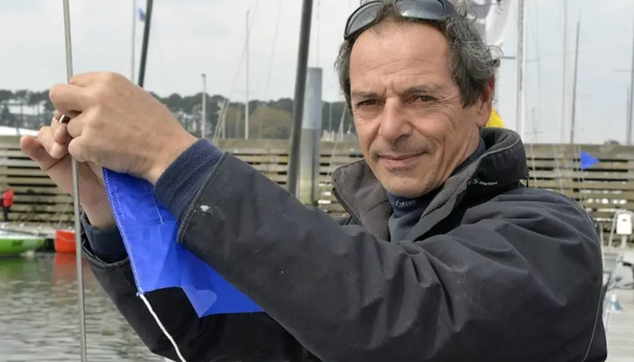 Marc Guillemot, skipper expérimenté, ajuste une voile bleue sur un ponton, prêt pour une nouvelle aventure en mer.