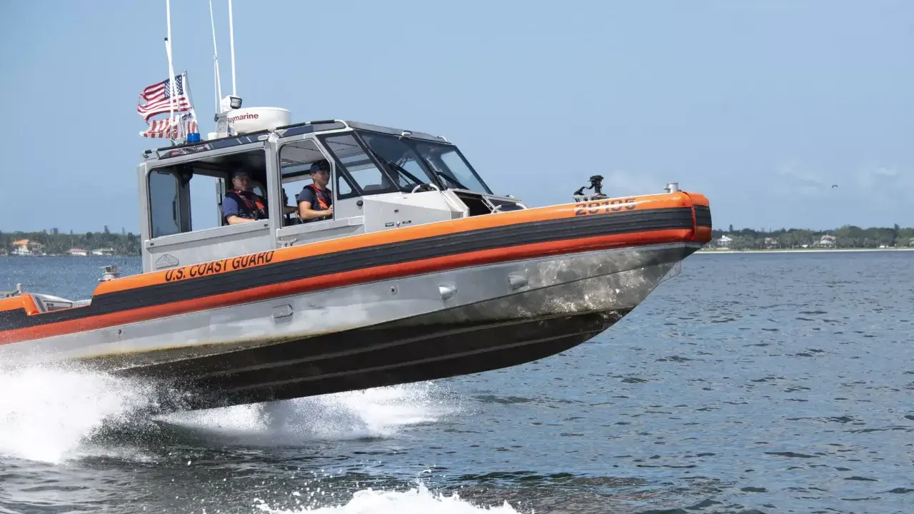Un bateau du garde côte américain file sur l'eau, créant une gerbe d'écume. Deux membres d'équipage sont visibles à l'intérieur.