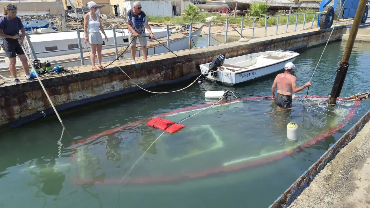 Un bateau accidenté est partiellement submergé dans un port. Des personnes sur le quai tentent de gérer la situation.