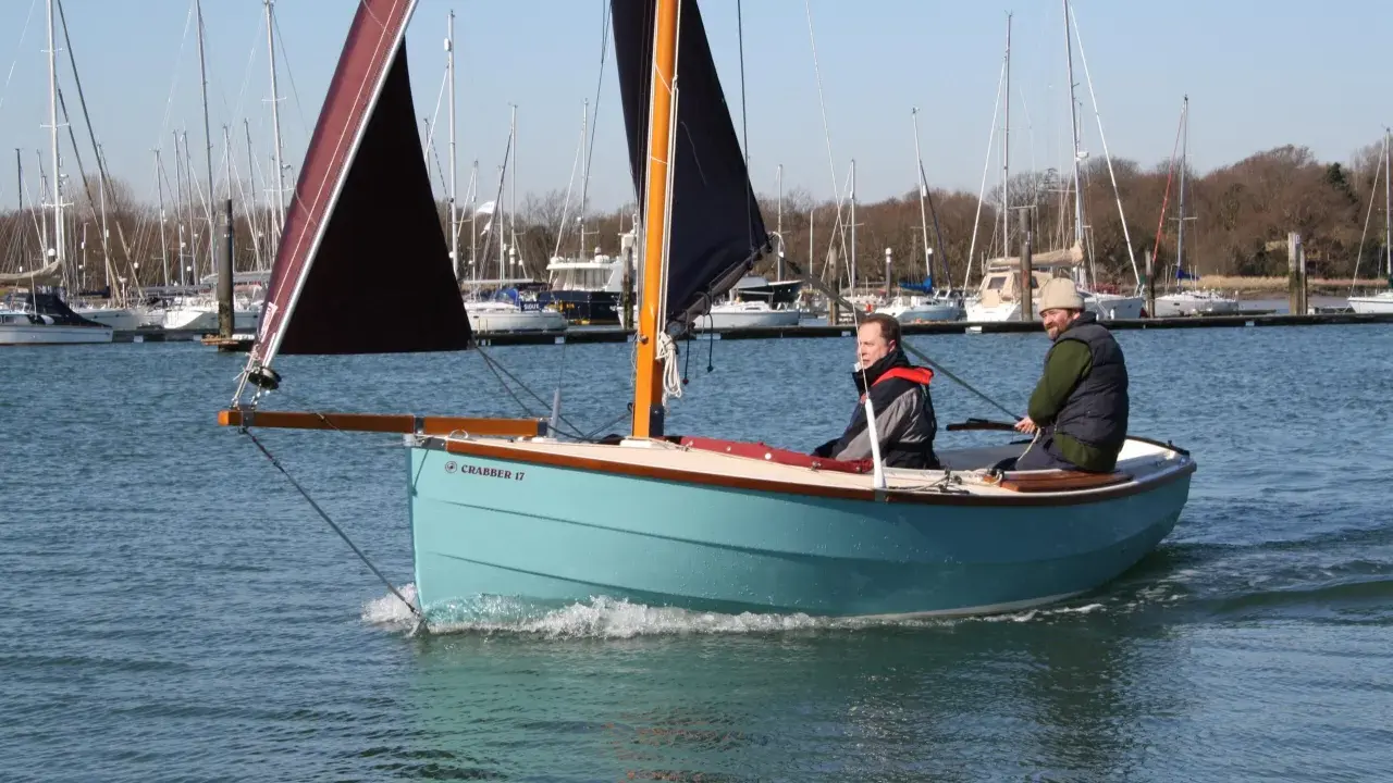 Deux hommes naviguent sur un petit bateau à voile bleu clair, avec d'autres voiliers en arrière-plan.