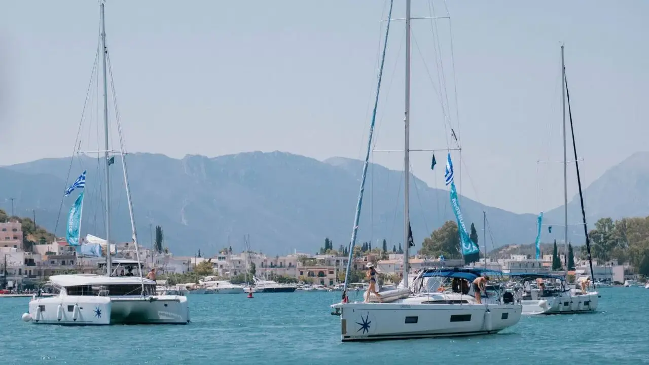 Trois voiliers naviguent dans une baie ensoleillée. Au premier plan, un catamaran monocoque blanc avec des voiles bleues et blanches. Derrière, un autre voilier blanc avec des personnes à bord. Au loin, une ville côtière et des montagnes.