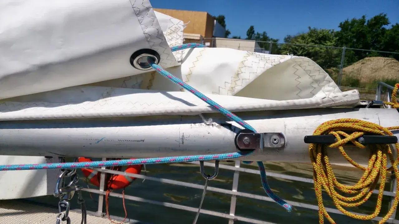 Le cordage bleu passe dans un œillet de la voile, prêt à ajuster la bosse de ris. Une amarre jaune est enroulée sur un taquet.