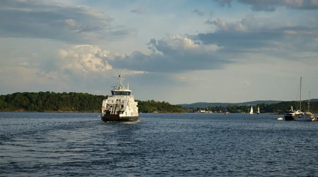 Un ferry blanc navigue sur une eau bleue, laissant une traînée derrière lui. La vitesse du bateau est visible dans le sillage. Au loin, des voiliers et une côte boisée.