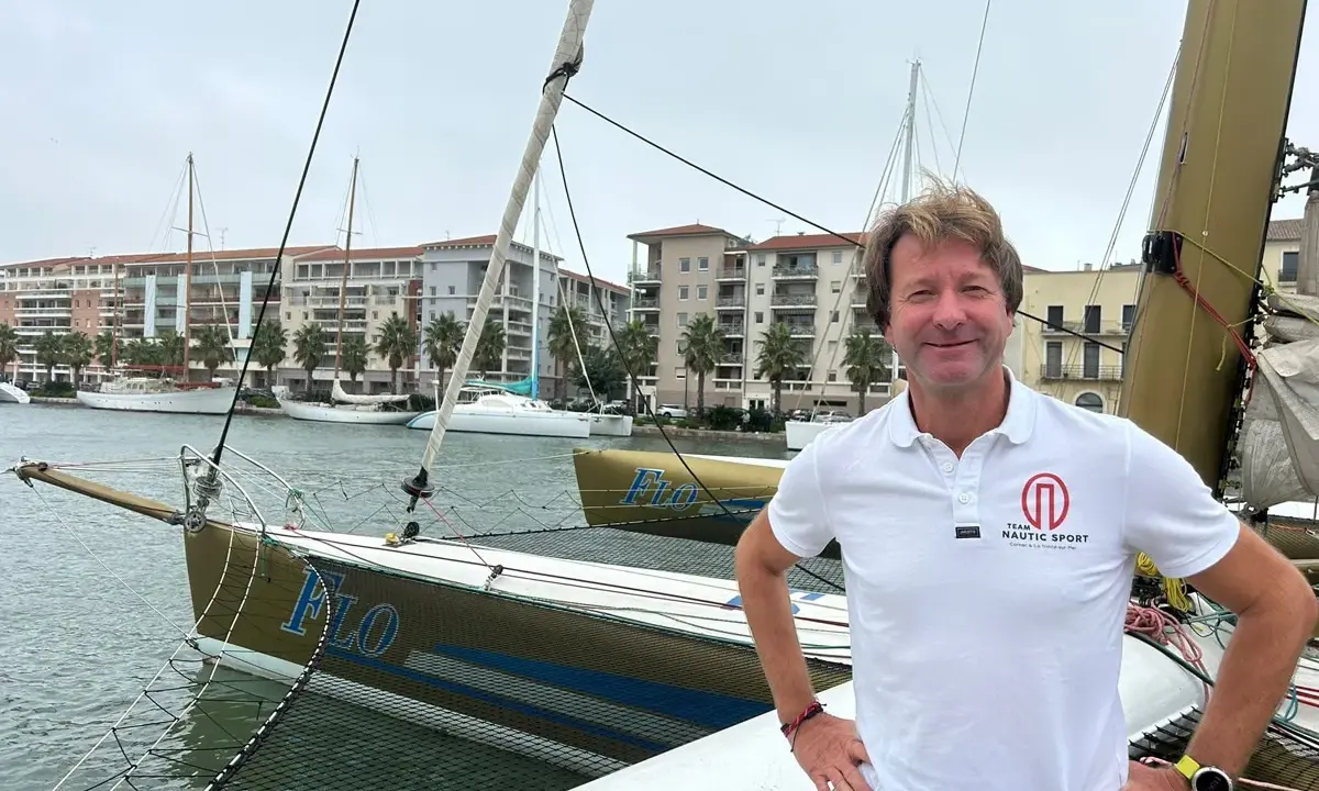 Un homme souriant sur un trimaran, peut-être le "Florence Arthaud", avec des bateaux et des immeubles en arrière-plan.