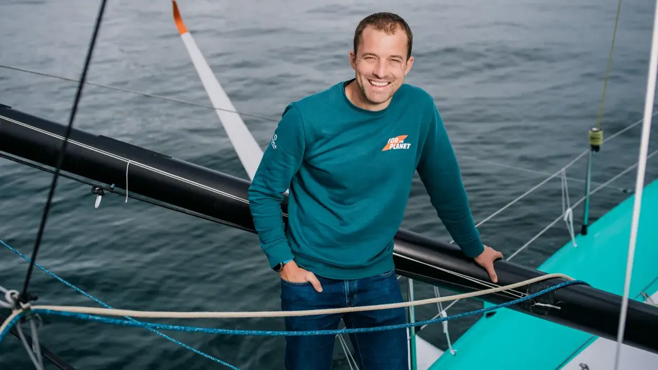 Sam Goodchild, souriant, sur un voilier, portant un sweat "For the Planet".