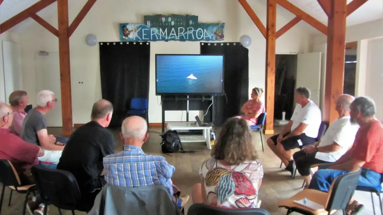 Gwen Chapalain, une femme assise, regarde un écran diffusant une image de bateau. Un groupe écoute attentivement.