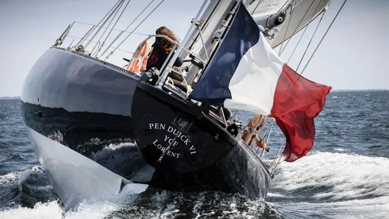 Le voilier Pen Duick VI fend les vagues, pavillon français au vent. Une silhouette se tient à la barre.