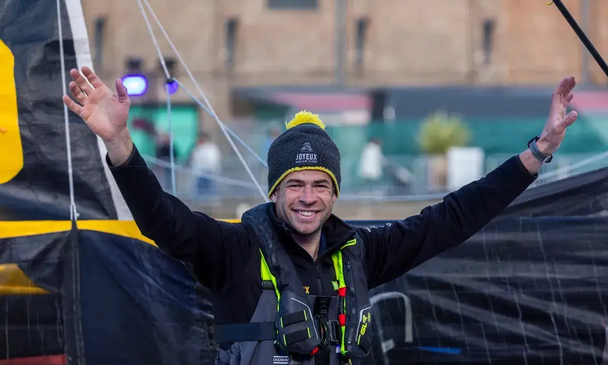Nicolas d'Estais, marin souriant, bras levés, porte un bonnet et un gilet de sauvetage, prêt pour l'aventure en mer.