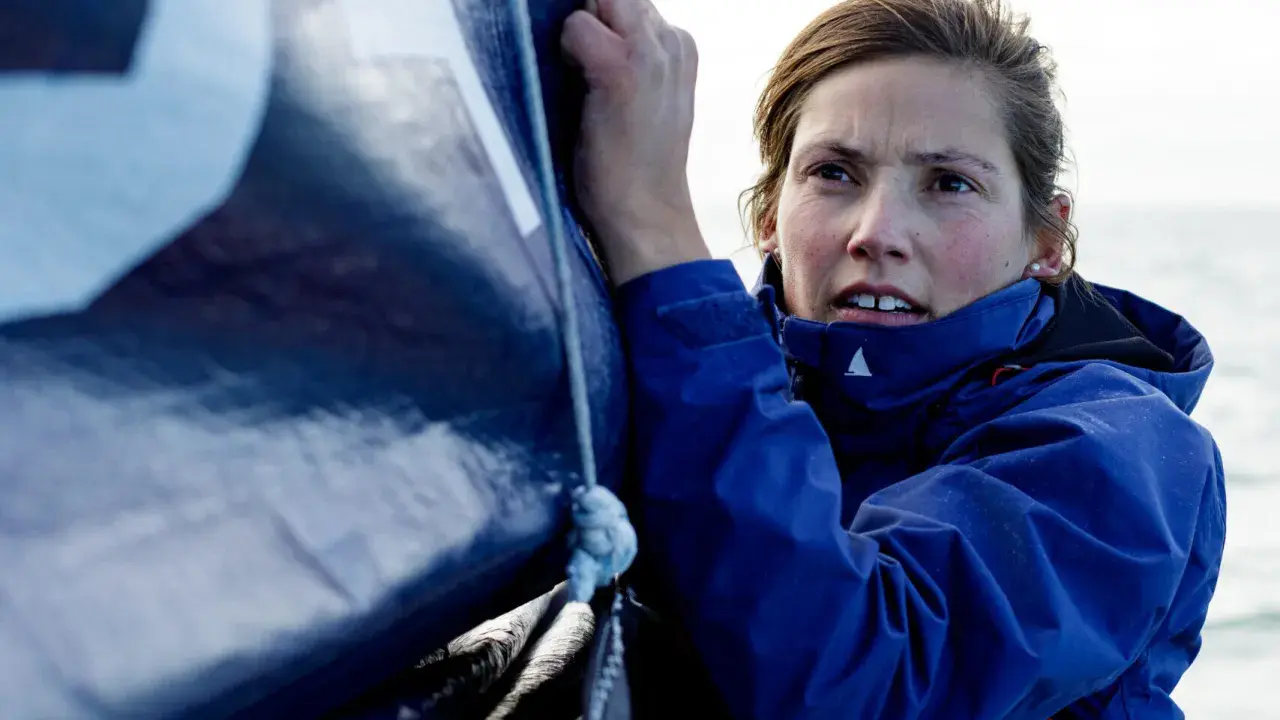 Tiphaine Ragueneau, concentrée, ajuste une voile bleue sur un bateau. La mer est visible en arrière-plan.