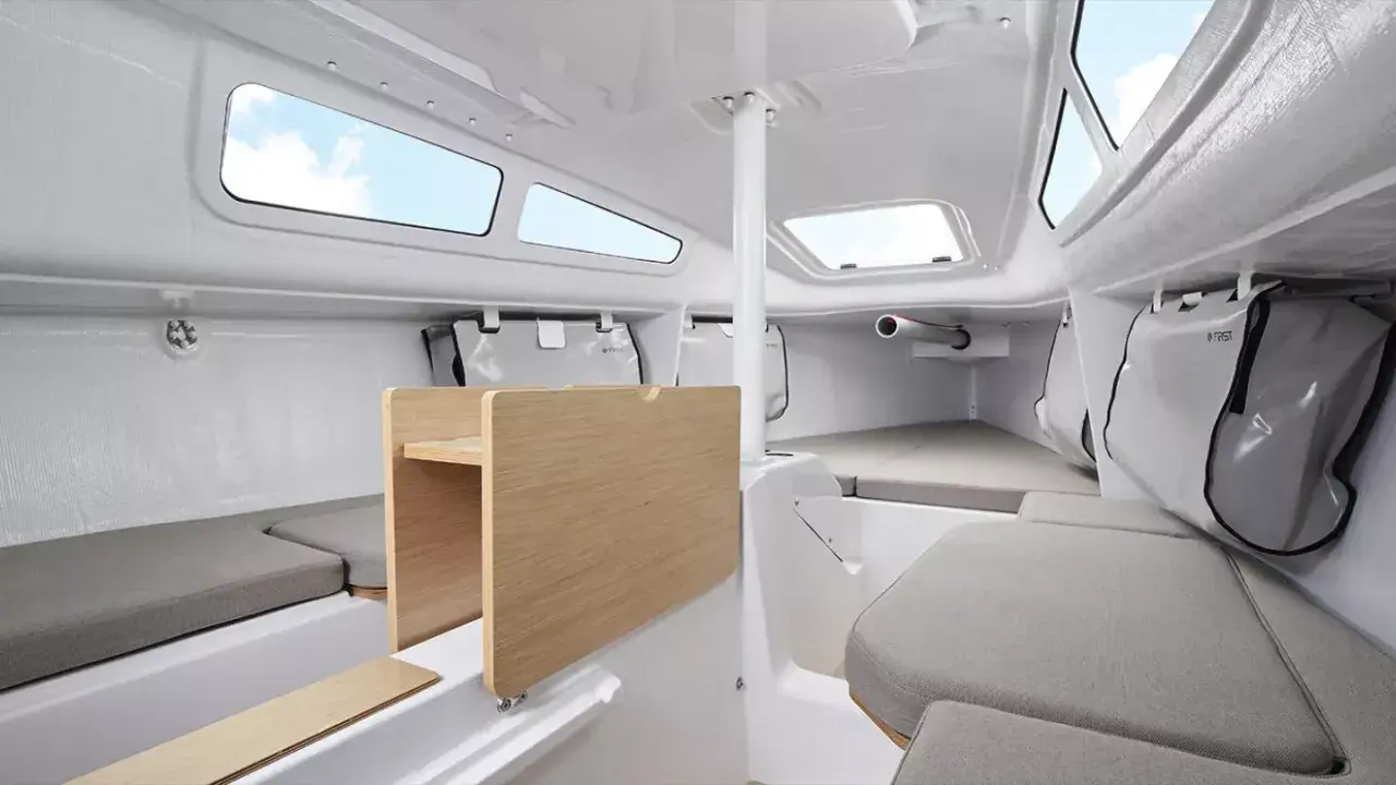 Intérieur du FIRST 24, cabine lumineuse avec banquettes grises, table en bois et hublots ouverts sur le ciel bleu.