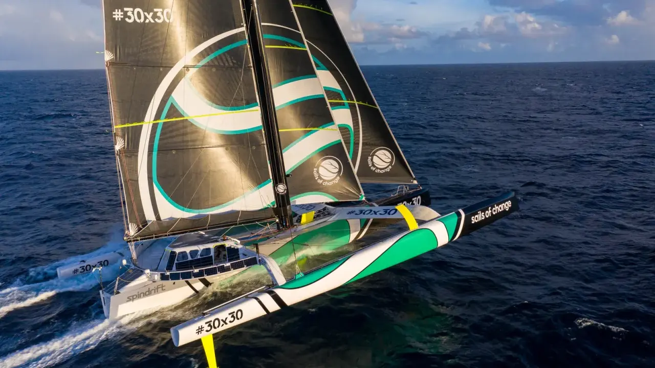 Un trimaran aux voiles noires et blanches, arborant le logo "sails of change", fend les vagues bleues sous un ciel nuageux.