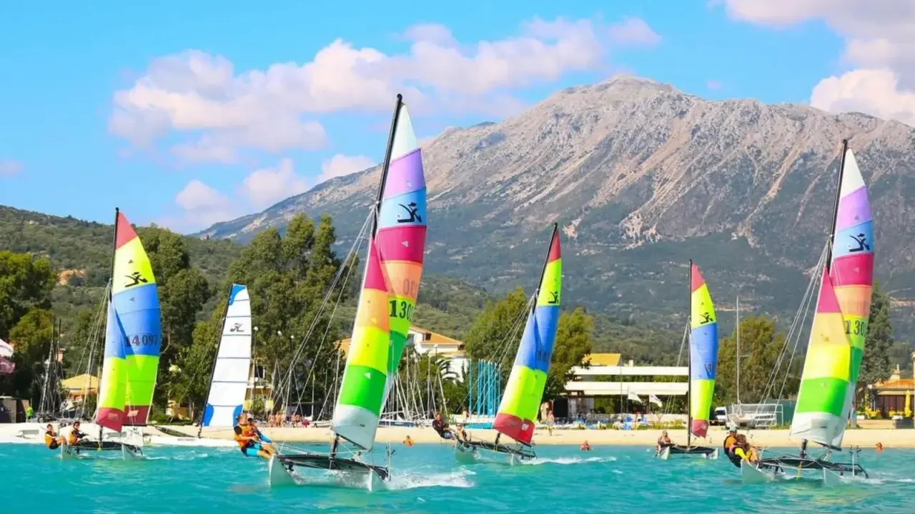 Plusieurs Hobie Cat 16 naviguent sur une mer turquoise, voiles multicolores, montagne en arrière-plan.